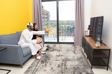 الاشارة باصبع السبابة الى شيء ما، الاصغاء والاستماع عبر الهيدفون، اللعب بألعاب الفيديو الترفيهية، استخدام الأجهزة التكنولوجية الحديثة، رجل عربي خليجي اماراتي يرتدي الكندورة والقبعة البيضاء يضع سماعات الرأس ويمسك بيده عصا التحكم يجلس مع زوجته في المنزل