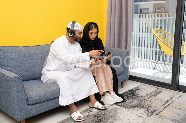 مفهوم المتعة والتسلية، اللعب بألعاب الفيديو الترفيهية، الاصغاء والاستماع عبر الهيدفون، استخدام الأجهزة التكنولوجية الحديثة، رجل عربي خليجي اماراتي يرتدي الكندورة والقبعة يضع سماعات الرأس ويمسك بيده عصا التحكم يجلس مع زوجته في المنزل، ايماءات التركيز والاندماج