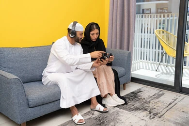 مفهوم المتعة والتسلية، اللعب بألعاب الفيديو الترفيهية، الاصغاء والاستماع عبر الهيدفون، استخدام الأجهزة التكنولوجية الحديثة، رجل عربي خليجي اماراتي يرتدي الكندورة والقبعة يضع سماعات الرأس ويمسك بيده عصا التحكم يجلس مع زوجته في المنزل، ايماءات التركيز والاندماج