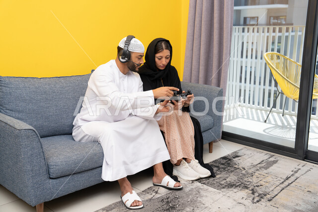 الاصغاء والاستماع عبر الهيدفون، اللعب بألعاب الفيديو الترفيهية، استخدام الأجهزة التكنولوجية الحديثة، رجل عربي خليجي اماراتي يرتدي الكندورة والقبعة البيضاء يضع سماعات الرأس ويمسك بيده عصا التحكم يجلس مع زوجته في المنزل، مفهوم التسلية والمتعة وقضاء وقت ممتع