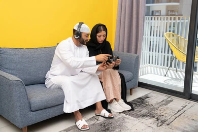 Listening and hearing through headphones, playing video games, using modern technological devices, an Emirati Gulf Arab man wearing a kandura and a white hat, wearing headphones and holding a joystick, sitting with his wife at home, the concept of entertainment, fun and spending a pleasant time