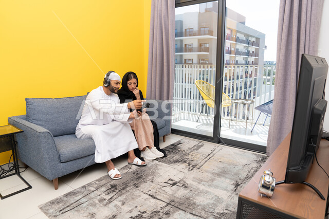 Listening and hearing through headphones, playing video games, using modern technological devices, an Emirati Gulf Arab man wearing a kandura and a white hat, wearing headphones and holding a joystick, sitting with his wife at home, the concept of entertainment, fun and spending a pleasant time