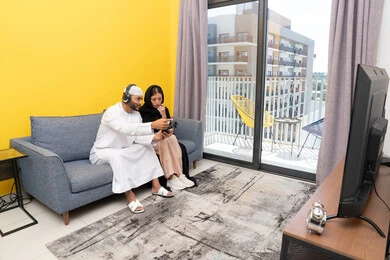 Listening and hearing through headphones, playing video games, using modern technological devices, an Emirati Gulf Arab man wearing a kandura and a white hat, wearing headphones and holding a joystick, sitting with his wife at home, the concept of entertainment, fun and spending a pleasant time