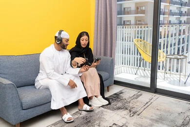 Listening and hearing through headphones, playing video games, using modern technological devices, an Emirati Gulf Arab man wearing a kandura and a white hat, wearing headphones and holding a joystick, sitting with his wife at home, the concept of entertainment, fun and spending a pleasant time