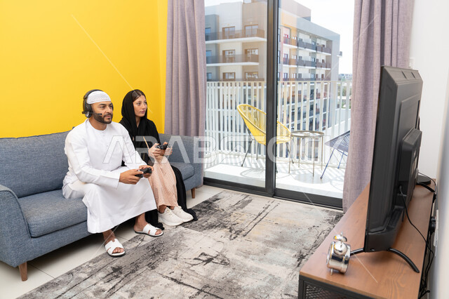 ايماءات التركيز والاندماج، مفهوم المتعة والتسلية، اللعب بألعاب الفيديو الترفيهية، الاصغاء والاستماع عبر الهيدفون، استخدام الأجهزة التكنولوجية الحديثة، رجل عربي خليجي اماراتي يرتدي الكندورة والقبعة يضع سماعات الرأس ويمسك بيده عصا التحكم يجلس مع زوجته في المنزل