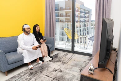 ايماءات التركيز والاندماج، مفهوم المتعة والتسلية، اللعب بألعاب الفيديو الترفيهية، الاصغاء والاستماع عبر الهيدفون، استخدام الأجهزة التكنولوجية الحديثة، رجل عربي خليجي اماراتي يرتدي الكندورة والقبعة يضع سماعات الرأس ويمسك بيده عصا التحكم يجلس مع زوجته في المنزل