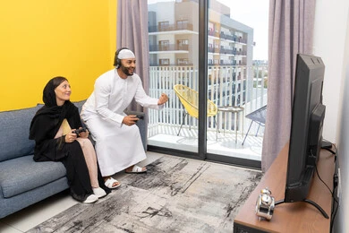 Playing video games, listening to headphones, using modern technological devices, an Emirati Gulf Arab man wearing a kandura and a white hat, wearing headphones and holding a joystick, sitting with his wife at home, raising his fist in gestures of enthusiasm and victory