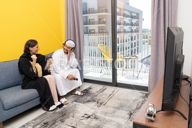 اللعب بألعاب الفيديو الترفيهية، الاصغاء والاستماع عبر الهيدفون، استخدام الأجهزة التكنولوجية الحديثة، رجل عربي خليجي اماراتي يرتدي الكندورة والقبعة البيضاء يضع سماعات الرأس ويمسك بيده عصا التحكم يجلس مع زوجته في المنزل، رفع قبضة اليد بايماءات الحماس والفوز