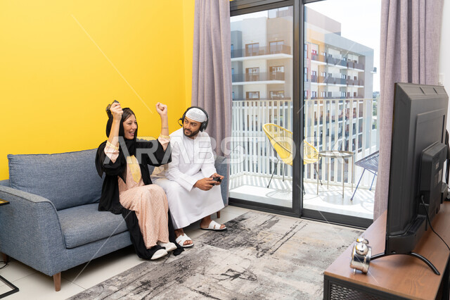 Raising a fist with gestures of enthusiasm and victory, playing entertaining video games, listening and paying attention through headphones, using modern technological devices, an Arab Gulf Emirati man wearing a kandura and a white hat, putting on headphones and holding a joystick, sitting with his wife at home