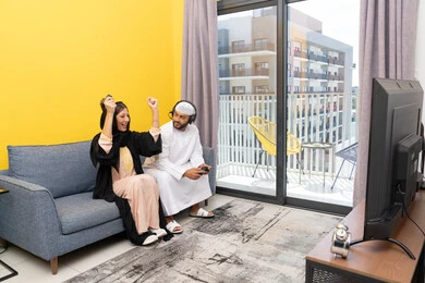 رفع قبضة اليد بايماءات الحماس والفوز، اللعب بألعاب الفيديو الترفيهية، الاستماع والاصغاء عبر الهيدفون، استخدام الأجهزة التكنولوجية الحديثة، رجل عربي خليجي اماراتي يرتدي الكندورة والقبعة البيضاء يضع سماعات الرأس ويمسك بيده عصا التحكم يجلس مع زوجته في المنزل