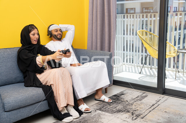Listening and hearing through headphones, playing video games, using modern technological devices, an Emirati Gulf Arab man wearing a kandura and a white hat, wearing headphones and holding a joystick, sitting with his wife at home, the concept of entertainment, fun and spending a pleasant time