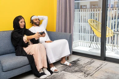 الاصغاء والاستماع عبر الهيدفون، اللعب بألعاب الفيديو الترفيهية، استخدام الأجهزة التكنولوجية الحديثة، رجل عربي خليجي اماراتي يرتدي الكندورة والقبعة البيضاء يضع سماعات الرأس ويمسك بيده عصا التحكم يجلس مع زوجته في المنزل، مفهوم التسلية والمتعة وقضاء وقت ممتع