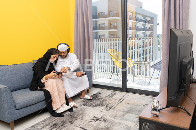 ايماءات التركيز والاندماج، مفهوم المتعة والتسلية، اللعب بألعاب الفيديو الترفيهية، الاصغاء والاستماع عبر الهيدفون، استخدام الأجهزة التكنولوجية الحديثة، رجل عربي خليجي اماراتي يرتدي الكندورة والقبعة يضع سماعات الرأس ويمسك بيده عصا التحكم يجلس مع زوجته في المنزل