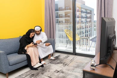 ايماءات التركيز والاندماج، مفهوم المتعة والتسلية، اللعب بألعاب الفيديو الترفيهية، الاصغاء والاستماع عبر الهيدفون، استخدام الأجهزة التكنولوجية الحديثة، رجل عربي خليجي اماراتي يرتدي الكندورة والقبعة يضع سماعات الرأس ويمسك بيده عصا التحكم يجلس مع زوجته في المنزل