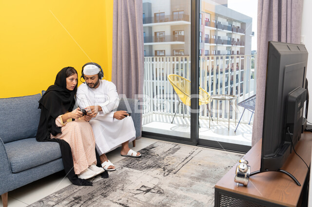 مفهوم المتعة والتسلية، اللعب بألعاب الفيديو الترفيهية، الاصغاء والاستماع عبر الهيدفون، استخدام الأجهزة التكنولوجية الحديثة، رجل عربي خليجي اماراتي يرتدي الكندورة والقبعة يضع سماعات الرأس ويمسك بيده عصا التحكم يجلس مع زوجته في المنزل، ايماءات التركيز والاندماج
