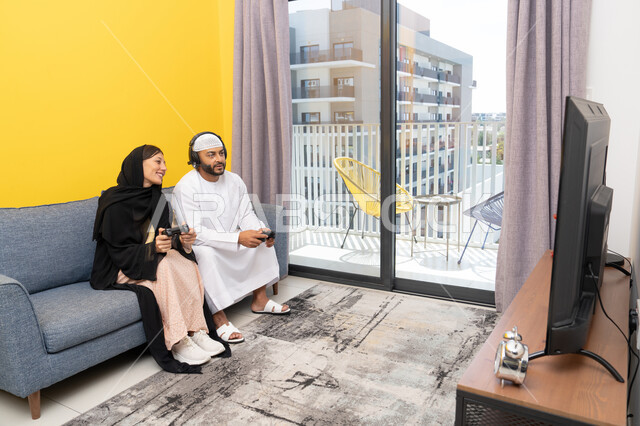 الاصغاء والاستماع عبر الهيدفون، اللعب بألعاب الفيديو الترفيهية، استخدام الأجهزة التكنولوجية الحديثة، رجل عربي خليجي اماراتي يرتدي الكندورة والقبعة البيضاء يضع سماعات الرأس ويمسك بيده عصا التحكم يجلس مع زوجته في المنزل، مفهوم التسلية والمتعة وقضاء وقت ممتع