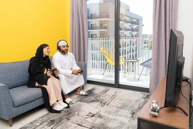 Listening and hearing through headphones, playing video games, using modern technological devices, an Emirati Gulf Arab man wearing a kandura and a white hat, wearing headphones and holding a joystick, sitting with his wife at home, the concept of entertainment, fun and spending a pleasant time