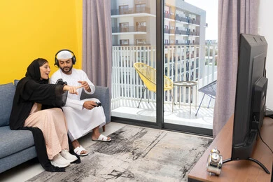 مفهوم المتعة والتسلية، اللعب بألعاب الفيديو الترفيهية، الاصغاء والاستماع عبر الهيدفون، استخدام الأجهزة التكنولوجية الحديثة، رجل عربي خليجي اماراتي يرتدي الكندورة والقبعة يضع سماعات الرأس ويمسك بيده عصا التحكم يجلس مع زوجته في المنزل، ايماءات التركيز والاندماج