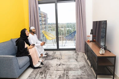 ايماءات التركيز والاندماج، مفهوم المتعة والتسلية، اللعب بألعاب الفيديو الترفيهية، الاصغاء والاستماع عبر الهيدفون، استخدام الأجهزة التكنولوجية الحديثة، رجل عربي خليجي اماراتي يرتدي الكندورة والقبعة يضع سماعات الرأس ويمسك بيده عصا التحكم يجلس مع زوجته في المنزل