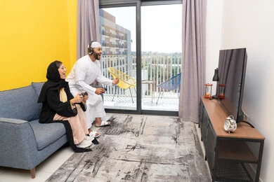 الاصغاء والاستماع عبر الهيدفون، اللعب بألعاب الفيديو الترفيهية، استخدام الأجهزة التكنولوجية الحديثة، رجل عربي خليجي اماراتي يرتدي الكندورة والقبعة البيضاء يضع سماعات الرأس ويمسك بيده عصا التحكم يجلس مع زوجته في المنزل، رفع قبضة اليد بايماءات الحماس والفوز