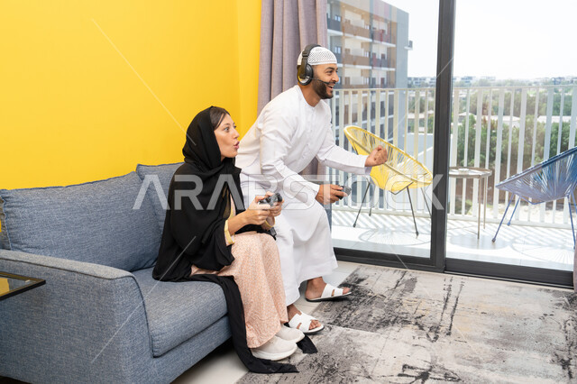 رفع قبضة اليد بايماءات الحماس والفوز، الاصغاء والاستماع عبر الهيدفون، اللعب بألعاب الفيديو الترفيهية، استخدام الأجهزة التكنولوجية الحديثة، رجل عربي خليجي اماراتي يرتدي الكندورة والقبعة البيضاء يضع سماعات الرأس ويمسك بيده عصا التحكم يجلس مع زوجته في المنزل