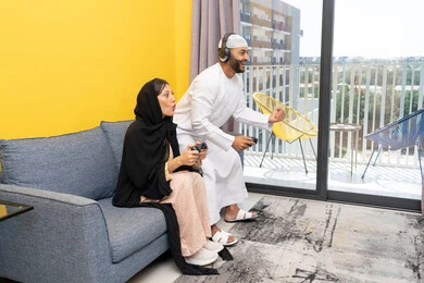 رفع قبضة اليد بايماءات الحماس والفوز، الاصغاء والاستماع عبر الهيدفون، اللعب بألعاب الفيديو الترفيهية، استخدام الأجهزة التكنولوجية الحديثة، رجل عربي خليجي اماراتي يرتدي الكندورة والقبعة البيضاء يضع سماعات الرأس ويمسك بيده عصا التحكم يجلس مع زوجته في المنزل