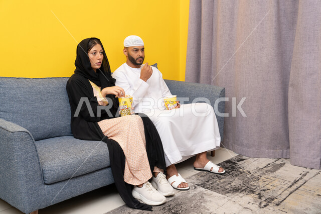 نشاط منزلي ترفيهي جماعي, المتعة في تناول الفشار، قضاء وقت ممتع بين الزوجان, رجل عربي خليجي اماراتي يرتدي الكندورة والقبعة البيضاء يجلس مع زوجته في المنزل، مشاهدة الافلام والعروض المسرحية بأيماءات السعادة و السرور، متابعة برامج التلفاز، الاستمتاع باوقات الفراغ