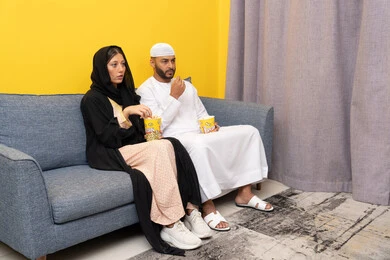 Group home entertainment activity, having fun eating popcorn, spending quality time between spouses, an Emirati Gulf Arab man wearing a kandura and a white hat sitting with his wife at home, watching movies and plays with gestures of happiness and pleasure, following TV programs, enjoying free time