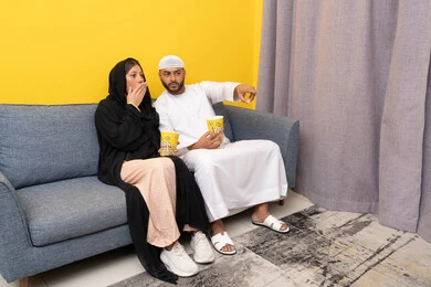 المتعة في تناول الفشار، قضاء وقت ممتع بين الزوجان, رجل عربي خليجي اماراتي يرتدي الكندورة والقبعة البيضاء يجلس مع زوجته في المنزل، مشاهدة الافلام والعروض المسرحية بأيماءات السعادة و السرور، متابعة برامج التلفاز، الاستمتاع باوقات الفراغ، نشاط منزلي ترفيهي جماعي