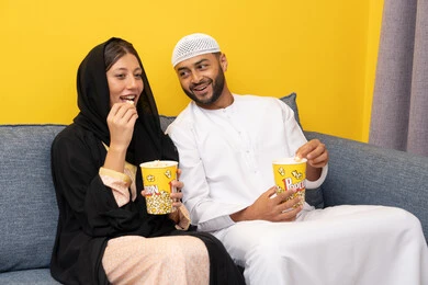 الاستمتاع باوقات الفراغ, نشاط منزلي ترفيهي جماعي, المتعة في تناول الفشار، قضاء وقت ممتع بين الزوجان, رجل عربي خليجي اماراتي يرتدي الكندورة والقبعة البيضاء يجلس مع زوجته في المنزل، مشاهدة الافلام والعروض المسرحية بأيماءات السعادة و السرور، متابعة برامج التلفاز