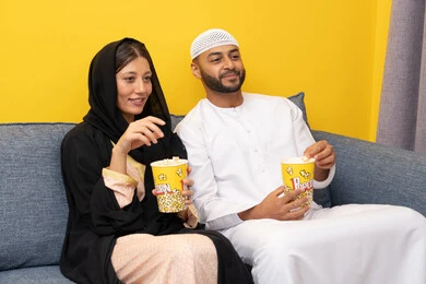 المتعة في تناول الفشار، قضاء وقت ممتع بين الزوجان, رجل عربي خليجي اماراتي يرتدي الكندورة والقبعة البيضاء يجلس مع زوجته في المنزل، مشاهدة الافلام والعروض المسرحية بأيماءات السعادة و السرور، متابعة برامج التلفاز، الاستمتاع باوقات الفراغ، نشاط منزلي ترفيهي جماعي