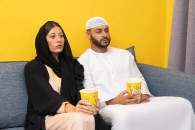 Enjoying free time, a group home entertainment activity, having fun eating popcorn, spending quality time between spouses, an Emirati Gulf Arab man wearing a kandura and a white hat sitting with his wife at home, watching movies and theatrical performances with gestures of happiness and pleasure, following TV programs