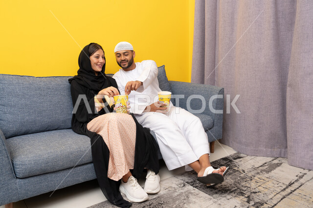 نشاط منزلي ترفيهي جماعي, المتعة في تناول الفشار، قضاء وقت ممتع بين الزوجان, رجل عربي خليجي اماراتي يرتدي الكندورة والقبعة البيضاء يجلس مع زوجته في المنزل، مشاهدة الافلام والعروض المسرحية بأيماءات السعادة و السرور، متابعة برامج التلفاز، الاستمتاع باوقات الفراغ