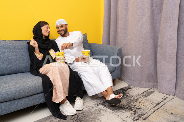 قضاء وقت ممتع بين الزوجان, رجل عربي خليجي اماراتي يرتدي الكندورة والقبعة البيضاء يجلس مع زوجته في المنزل، مشاهدة الافلام والعروض المسرحية بأيماءات السعادة و السرور، متابعة برامج التلفاز، الاستمتاع باوقات الفراغ، نشاط منزلي ترفيهي جماعي، المتعة في تناول الفشار