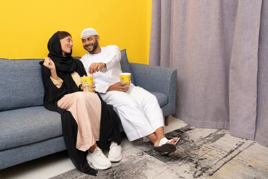 قضاء وقت ممتع بين الزوجان, رجل عربي خليجي اماراتي يرتدي الكندورة والقبعة البيضاء يجلس مع زوجته في المنزل، مشاهدة الافلام والعروض المسرحية بأيماءات السعادة و السرور، متابعة برامج التلفاز، الاستمتاع باوقات الفراغ، نشاط منزلي ترفيهي جماعي، المتعة في تناول الفشار