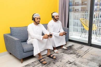 المنافسة والمشاركة بالألعاب، مفهوم المتعة والتسلية، نشاط شبابي ترفيهي، اللعب بألعاب الفيديو الترفيهية مع الأصدقاء في المنزل، الاصغاء والاستماع عبر الهيدفون، رجلان عربيان خليجيان اماراتيان يرتديان الكندورة والقبعة يضعان سماعات الرأس ويمسكان بيدهما عصا التحكم