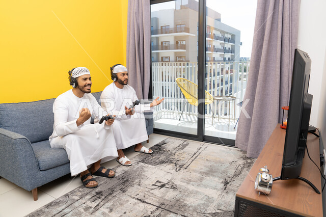 المنافسة والمشاركة بالألعاب، مفهوم المتعة والتسلية، نشاط شبابي ترفيهي، اللعب بألعاب الفيديو الترفيهية مع الأصدقاء في المنزل، الاصغاء والاستماع عبر الهيدفون، رجلان عربيان خليجيان اماراتيان يرتديان الكندورة والقبعة يضعان سماعات الرأس ويمسكان بيدهما عصا التحكم