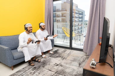 المنافسة والمشاركة بالألعاب، مفهوم المتعة والتسلية، نشاط شبابي ترفيهي، اللعب بألعاب الفيديو الترفيهية مع الأصدقاء في المنزل، الاصغاء والاستماع عبر الهيدفون، رجلان عربيان خليجيان اماراتيان يرتديان الكندورة والقبعة يضعان سماعات الرأس ويمسكان بيدهما عصا التحكم