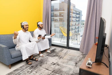 اللعب بألعاب الفيديو الترفيهية مع الأصدقاء في المنزل، الاصغاء والاستماع عبر الهيدفون، رجلان عربيان خليجيان اماراتيان يرتديان الكندورة والقبعة يضعان سماعات الرأس ويمسكان بيدهما عصا التحكم، المنافسة والمشاركة بالألعاب، مفهوم المتعة والتسلية، نشاط شبابي ترفيهي
