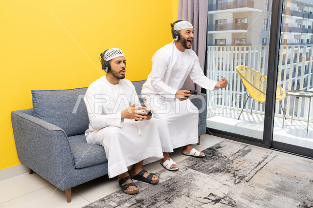 نشاط شبابي ترفيهي، مفهوم المتعة والتسلية، اللعب بألعاب الفيديو الترفيهية مع الأصدقاء في المنزل، الاصغاء والاستماع عبر الهيدفون، استخدام الأجهزة التكنولوجية الحديثة، رجلان عربيان خليجيان اماراتيان يرتديان الكندورة والقبعة يضعان سماعات الرأس ويمسكان بيدهما عصا التحكم