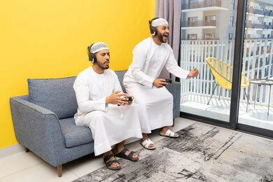 نشاط شبابي ترفيهي، مفهوم المتعة والتسلية، اللعب بألعاب الفيديو الترفيهية مع الأصدقاء في المنزل، الاصغاء والاستماع عبر الهيدفون، استخدام الأجهزة التكنولوجية الحديثة، رجلان عربيان خليجيان اماراتيان يرتديان الكندورة والقبعة يضعان سماعات الرأس ويمسكان بيدهما عصا التحكم