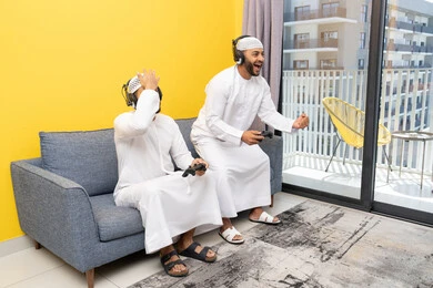 نشاط شبابي ترفيهي، مفهوم المتعة والتسلية، اللعب بألعاب الفيديو الترفيهية مع الأصدقاء في المنزل، الاصغاء والاستماع عبر الهيدفون، استخدام الأجهزة التكنولوجية الحديثة، رجلان عربيان خليجيان اماراتيان يرتديان الكندورة والقبعة يضعان سماعات الرأس ويمسكان بيدهما عصا التحكم