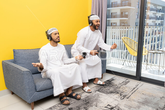نشاط شبابي ترفيهي، مفهوم المتعة والتسلية، اللعب بألعاب الفيديو الترفيهية مع الأصدقاء في المنزل، الاصغاء والاستماع عبر الهيدفون، استخدام الأجهزة التكنولوجية الحديثة، رجلان عربيان خليجيان اماراتيان يرتديان الكندورة والقبعة يضعان سماعات الرأس ويمسكان بيدهما عصا التحكم