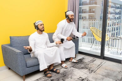 نشاط شبابي ترفيهي، مفهوم المتعة والتسلية، اللعب بألعاب الفيديو الترفيهية مع الأصدقاء في المنزل، الاصغاء والاستماع عبر الهيدفون، استخدام الأجهزة التكنولوجية الحديثة، رجلان عربيان خليجيان اماراتيان يرتديان الكندورة والقبعة يضعان سماعات الرأس ويمسكان بيدهما عصا التحكم