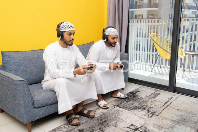 Competition and participation in games, the concept of fun and entertainment, a recreational youth activity, playing recreational video games with friends at home, listening and hearing through headphones, two Emirati Gulf Arab men wearing kandoras and hats, putting on headphones and holding joysticks