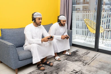 المنافسة والمشاركة بالألعاب، مفهوم المتعة والتسلية، نشاط شبابي ترفيهي، اللعب بألعاب الفيديو الترفيهية مع الأصدقاء في المنزل، الاصغاء والاستماع عبر الهيدفون، رجلان عربيان خليجيان اماراتيان يرتديان الكندورة والقبعة يضعان سماعات الرأس ويمسكان بيدهما عصا التحكم