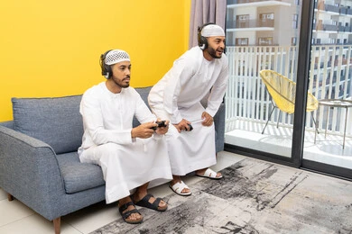 Recreational youth activity, the concept of fun and entertainment, playing recreational video games with friends at home, listening and hearing through headphones, using modern technological devices, two Emirati Gulf Arab men wearing kandoras and hats, putting on headphones and holding joysticks