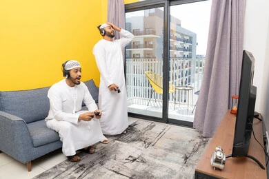 المنافسة والمشاركة بالألعاب، مفهوم المتعة والتسلية، نشاط شبابي ترفيهي، اللعب بألعاب الفيديو الترفيهية مع الأصدقاء في المنزل، الاصغاء والاستماع عبر الهيدفون، رجلان عربيان خليجيان اماراتيان يرتديان الكندورة والقبعة يضعان سماعات الرأس ويمسكان بيدهما عصا التحكم