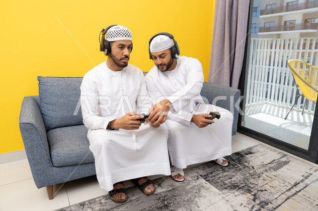 اللعب بألعاب الفيديو الترفيهية مع الأصدقاء في المنزل، الاصغاء والاستماع عبر الهيدفون، رجلان عربيان خليجيان اماراتيان يرتديان الكندورة والقبعة يضعان سماعات الرأس ويمسكان بيدهما عصا التحكم، المنافسة والمشاركة بالألعاب، مفهوم المتعة والتسلية، نشاط شبابي ترفيهي