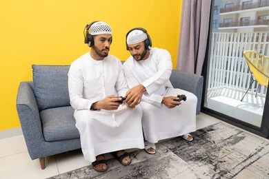 اللعب بألعاب الفيديو الترفيهية مع الأصدقاء في المنزل، الاصغاء والاستماع عبر الهيدفون، رجلان عربيان خليجيان اماراتيان يرتديان الكندورة والقبعة يضعان سماعات الرأس ويمسكان بيدهما عصا التحكم، المنافسة والمشاركة بالألعاب، مفهوم المتعة والتسلية، نشاط شبابي ترفيهي